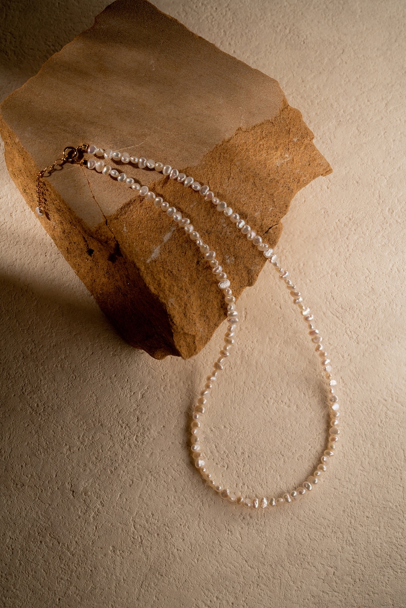 May include: A white pearl necklace with a gold clasp, laying on a brown rock.