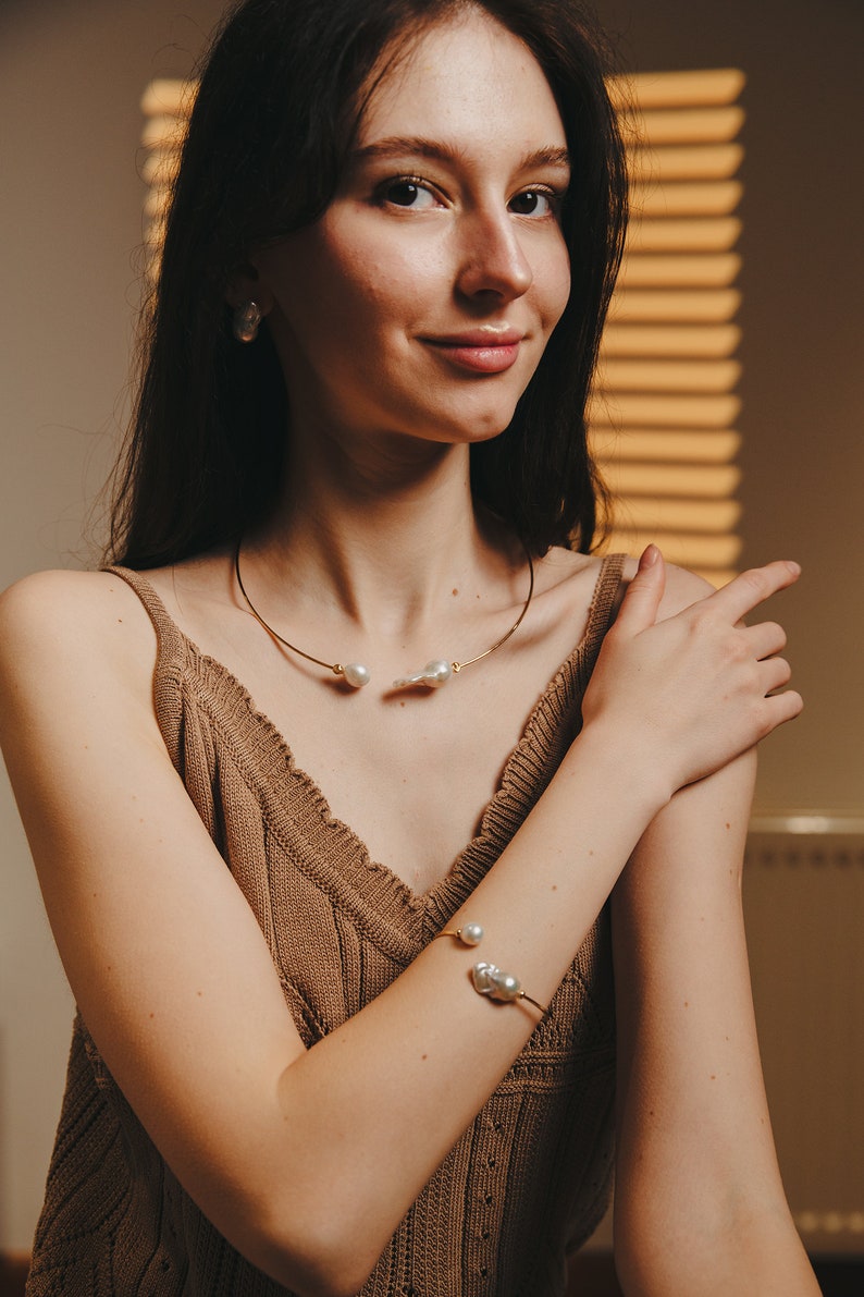 May include: A woman wearing a brown knitted tank top with a gold necklace and bracelet featuring pearl accents. The necklace is a thin choker style and the bracelet is a cuff style.