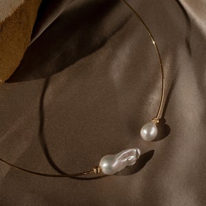 May include: A gold choker necklace with two large, irregular-shaped pearls. The necklace is on a brown fabric background.