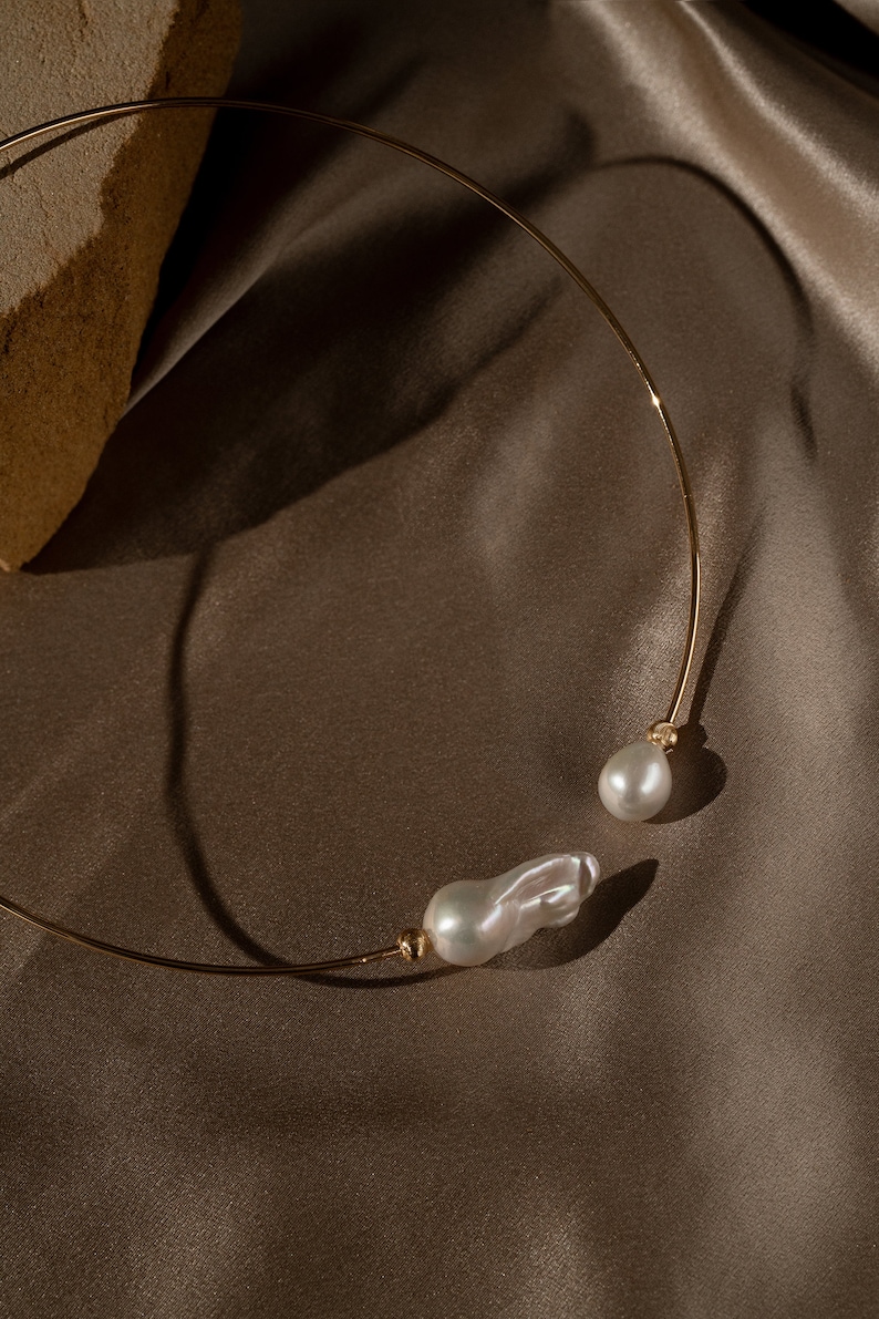 May include: A gold choker necklace with two large, irregular-shaped pearls. The necklace is on a brown fabric background.
