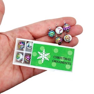 May include: A hand holds a white box containing miniature Christmas ornaments. The box has a green lid with a snowflake and the words "CHRISTMAS ORNAMENTS." The ornaments are colourful, with various designs and patterns.
