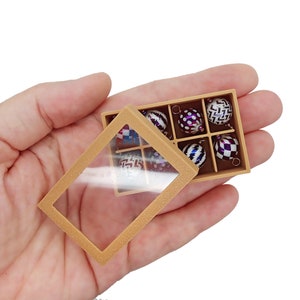 May include: A gold box with a clear lid, containing eight small, colorful Christmas ornaments with geometric patterns. The ornaments are arranged in a grid pattern inside the box.