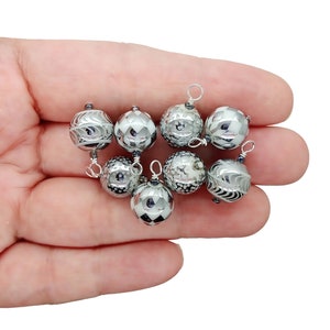 May include: A collection of eight silver-toned, spherical beads with intricate designs. Each bead has a small loop at the top, suggesting they are pendants or charms. The beads are held in a hand against a white background.