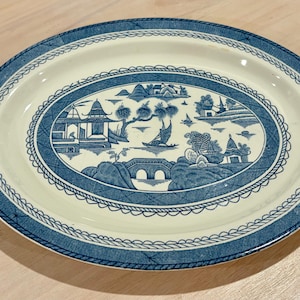 May include: An oval ceramic platter featuring a blue and white transferware pattern. The central scene depicts a landscape with pagodas, a bridge, and a boat on water, surrounded by a decorative border of rope and cross-hatch motifs.