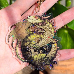 Könnte beinhalten: Ein Drachen-Schlüsselanhänger mit einem goldfarbenen Verschluss. Der Drache ist grün, braun und gelb mit roten Augen und einem detaillierten Flügeldesign. Der Schlüsselanhänger hat einen transparenten, glitzernden Rand.