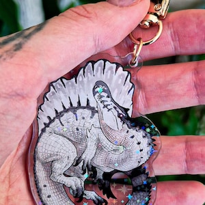 Op de afbeelding: Een doorzichtige acryl sleutelhanger met een gedetailleerde illustratie van een Spinosaurus dinosaurus in grijze en witte tinten. De dinosaurus is in een cirkelvormige houding gekruld met een opvallend rugzeil. Het oppervlak heeft een holografische glitterafwerking.