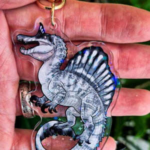 Op de afbeelding: Een doorzichtige acryl sleutelhanger met een gedetailleerde illustratie van een Spinosaurus dinosaurus in grijze en witte tinten. De dinosaurus heeft een opvallend rugzeil en is omringd door holografische sterren op het transparante oppervlak.