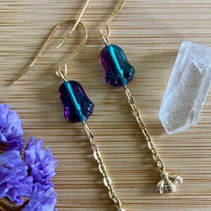 May include: A pair of gold-tone drop earrings featuring a purple and teal iridescent bell-shaped glass bead suspended from a gold-tone chain, ending in a small gold-tone bee charm. The earrings have gold-tone ear wires for pierced ears.