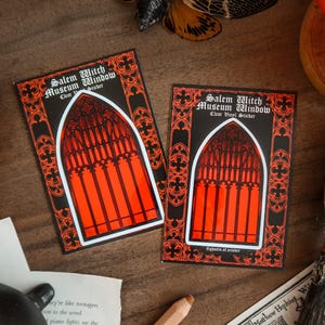 May include: Two rectangular stickers with a black border and a gothic window design in orange and red. The text "Salem Witch Museum Window" is at the top. The stickers are on a wooden surface.