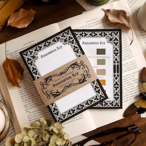 May include: Two black and white "Annotation Key" cards with decorative borders and a matching tag, displayed on open book pages. The tag reads "Ghosts of October" and features ornate scrollwork. Dried leaves and a candle holder complete the scene.