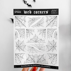 May include: A sheet of planner stickers with black and white spider web corner designs. The text "WEB CORNERS" and "planner stickers" are at the top. The sheet is labelled "handmade by @ghosts.of.october". Black bat decorations are in the background.