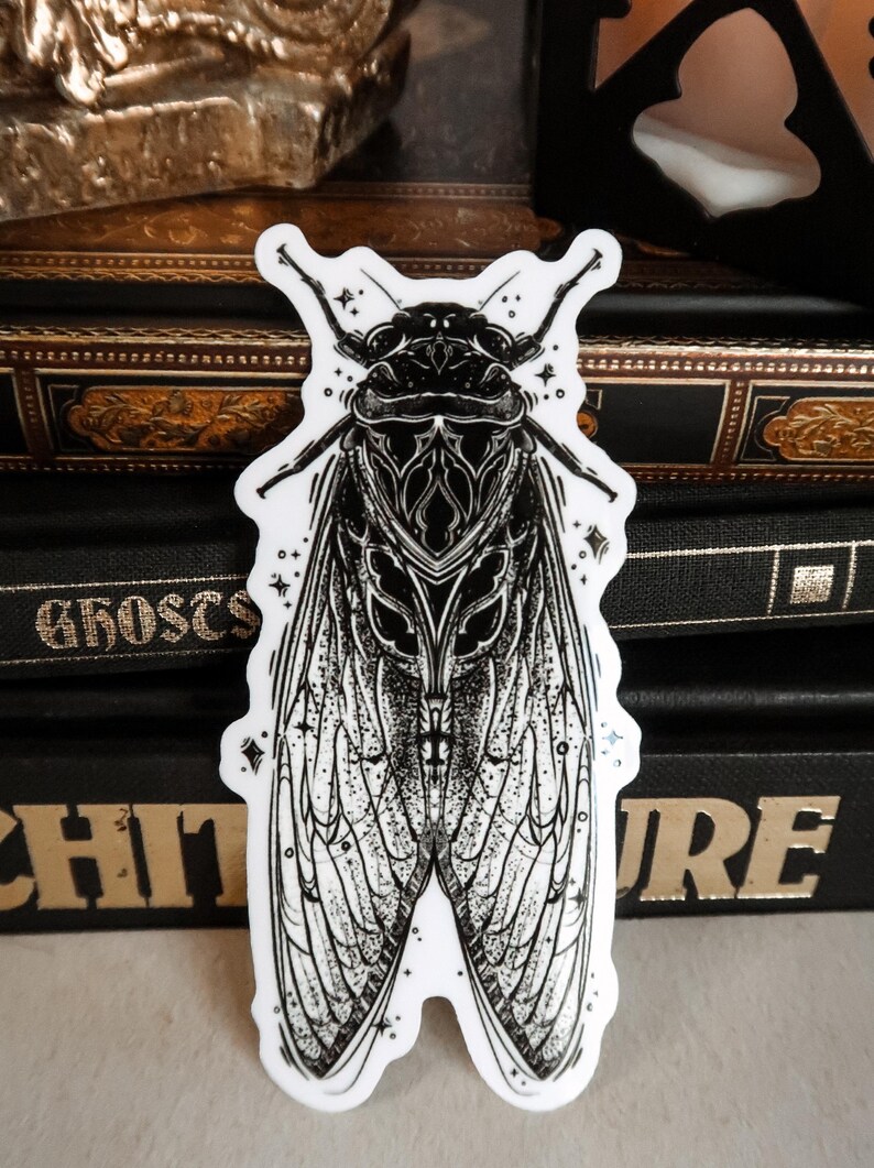 May include: A black and white sticker featuring a detailed illustration of a cicada. The insect has intricate wing patterns and is surrounded by small star-shaped accents. The sticker is on a stack of dark books.