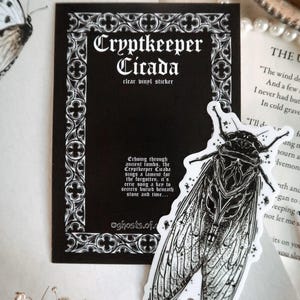 May include: A black and white clear vinyl sticker featuring a detailed illustration of a cicada insect. The sticker is next to a black card with the text "Cryptkeeper Cicada" and decorative border. The card also includes additional text.