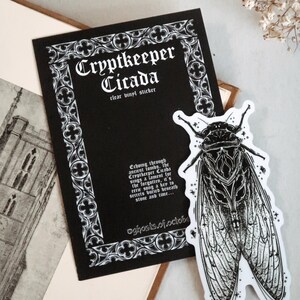 May include: A black and white clear vinyl sticker featuring a detailed cicada illustration. The sticker is next to a black card with the text "Cryptkeeper Cicada" and additional text. The design has a gothic aesthetic.