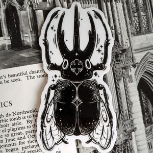 May include: Black and white sticker of a beetle with intricate details and a starry background. The beetle has large horns and wings.
