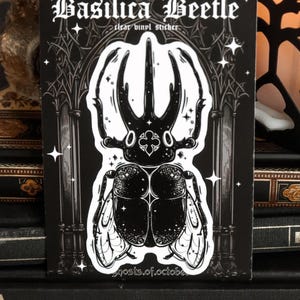 May include: Black and white sticker of a beetle with a gothic archway background. The sticker says "Basilica Beetle" and "clear vinyl sticker".