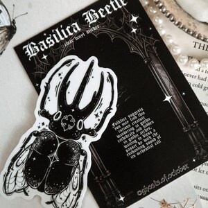 May include: A black and white sticker of a beetle with intricate details. The sticker is on a black background with white text that reads "Basilica Beetle clear vinyl sticker" and "Folklore suggests you may find this curious creature wandering in gothic cathedrals and hidden in gothic arches, waiting to bless a passer by with the enchanting sound of its orchestral call" and "oghosts.of.october".
