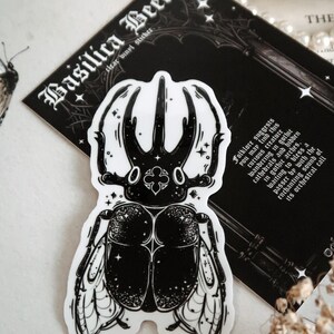 May include: Black and white sticker of a beetle with intricate details and a starry background. The sticker is labeled "Basilica Beetle" and has text about folklore and the creature's connection to gothic architecture.