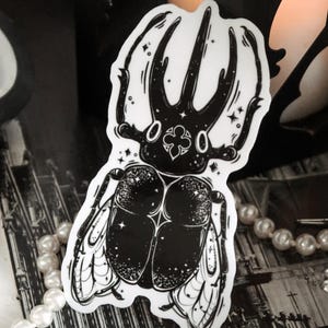 May include: Black and white sticker of a beetle with wings. The beetle has a detailed design with a star pattern and a clover shape on its head.