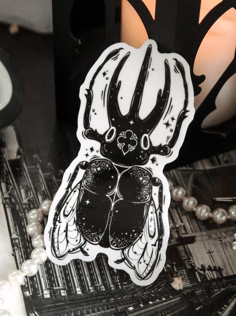 May include: Black and white sticker of a beetle with wings. The beetle has a detailed design with a star pattern and a clover shape on its head.