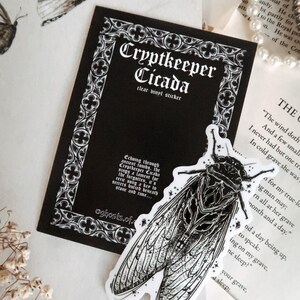 May include: A clear vinyl sticker featuring a detailed black and white illustration of a cicada insect. The sticker is labeled "Cryptkeeper Cicada" with gothic-style text on a black background. The design has a vintage, spooky aesthetic.