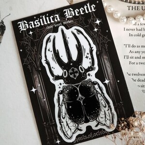 May include: Black and white sticker of a beetle with large horns and wings. The sticker is on a black background with a gothic archway and stars. The text "Basilica Beetle" and "clear vinyl sticker" are at the top of the sticker. The text "ghosts.of.october" is at the bottom of the sticker.