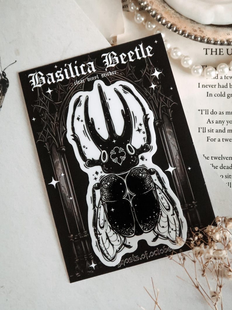 May include: Black and white sticker of a beetle with large horns and wings. The sticker is on a black background with a gothic archway and stars. The text "Basilica Beetle" and "clear vinyl sticker" are at the top of the sticker. The text "ghosts.of.october" is at the bottom of the sticker.