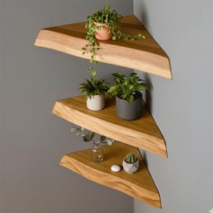 May include: Three wooden corner shelves, triangular in shape, with a natural wood grain. Each shelf displays small potted plants, including trailing vines and succulents. The shelves are mounted on a grey wall. The shelves are made of wood.