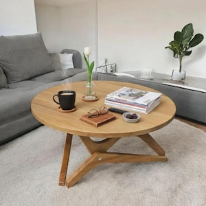 May include: A round, light-colored wooden coffee table with a three-legged base. A black mug of coffee sits on a coaster. Magazines, a small dish of chocolates, and a pair of glasses rest on the table. A vase with a white flower is also present.