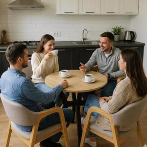 May include: A group of four people seated around a round wooden table, each holding a white coffee cup. The table is set in a modern kitchen with a light wood floor and neutral-toned cabinets. The chairs have light-colored upholstery.