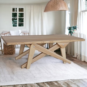 May include: A light wood dining table with a rectangular top and a unique X-shaped base. The table is made of natural wood with a light brown finish. The table is sitting on a white rug in a room with white walls and a window.