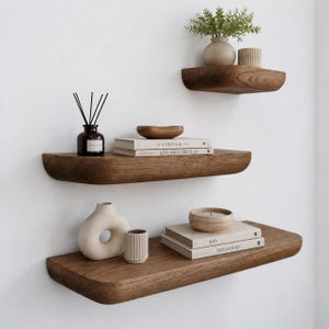 May include: Three floating wall shelves made of dark brown wood with rounded edges. The shelves hold decorative items including a small vase with greenery, a reed diffuser, books titled The Kinfolk and This Is Home, and ceramic bowls.