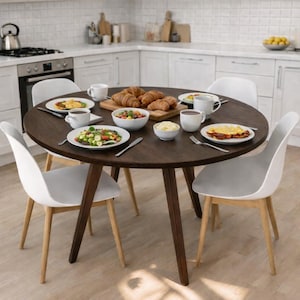 Op de afbeelding: Een ronde, donkerbruine houten eettafel, omringd door vier witte stoelen. De tafel is gedekt met borden met eten, kommen, kopjes en croissants. De stoelen hebben licht houten poten en witte zittingen.