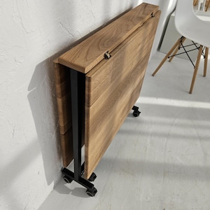 May include: A foldable table with a natural wood grain finish and a black metal frame. The table is shown in a collapsed, upright position, featuring hinged panels and small black caster wheels at the base for mobility and compact storage.