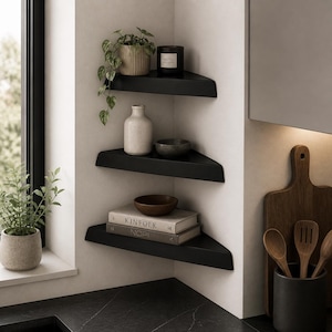 Peut inclure: Trois étagères d'angle noires avec divers objets décoratifs. L'étagère supérieure présente une plante en pot et une bougie noire avec l'inscription "KOTO". L'étagère du milieu contient un vase blanc et un petit bol gris. L'étagère inférieure expose des livres et un bol en bois.