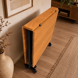 Op de afbeelding: Een lichtbruine, opvouwbare houten tafel met een zwart metalen frame en wielen. De tafel is gedeeltelijk opgevouwen, met de houtnerf zichtbaar. De tafel is ongeveer 76 cm lang wanneer opgevouwen.