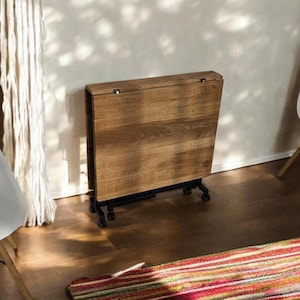 Puede incluir: Una mesa de madera plegable con un acabado de vetas de madera natural. La mesa es marrón y tiene una estructura de metal negro con ruedas. La mesa está parcialmente plegada y se apoya contra una pared blanca.