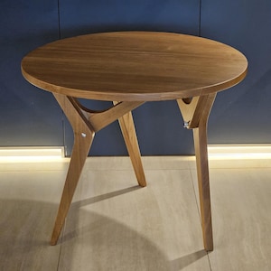 May include: A round wooden side table with a light brown finish. The table has three angled legs and a unique support structure. The table's top is approximately 24 inches in diameter.