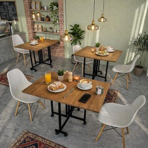May include: A cafe interior featuring several square wooden tables with black metal legs and white chairs. Each table is set with food and drinks, including croissants, coffee, and juice. Overhead, there are hanging lights, and the decor includes plants and small rugs.