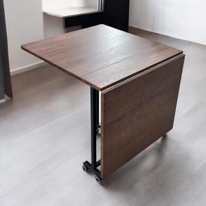 May include: A brown wooden folding table with black metal legs and wheels. The table is in a folded position, with one side of the table top folded up against the other.