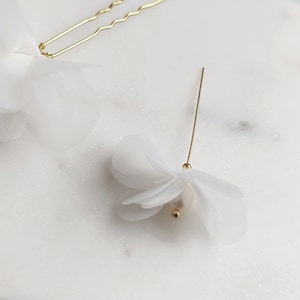 May include: Two white fabric flower hair clips. One clip is a single flower with a pearl center and a gold pin. The other clip is a cluster of smaller flowers with a gold bobby pin.