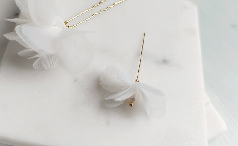 May include: Two white fabric flower hair clips. One clip is a single flower with a pearl center and a gold pin. The other clip is a cluster of smaller flowers with a gold bobby pin.