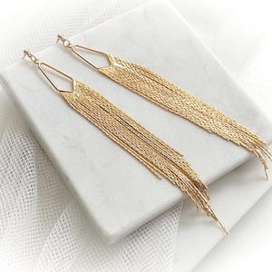 Könnte beinhalten: Ein Paar goldfarbene Hängeohrringe. Jeder Ohrring hat eine geometrische Oberseite mit einem langen, kaskadierenden Fransen aus filigranen Ketten. Die Ohrringe werden auf einer weißen Oberfläche präsentiert.