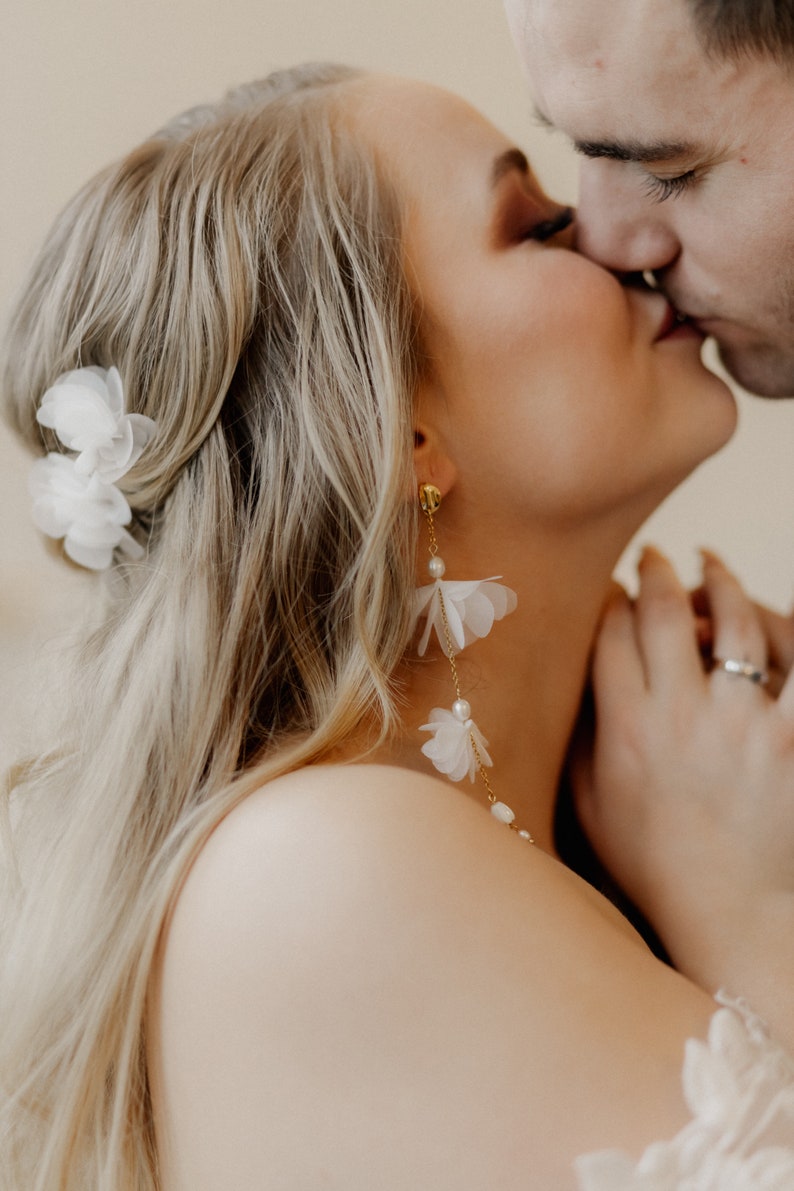 May include: A close-up of a bride and groom kissing. The bride is wearing a white flower in her hair and long, delicate white flower earrings.