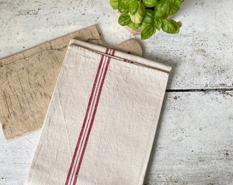 May include: Two rustic wooden cutting boards with a white linen tea towel with red stripes. The towel is folded and placed on top of the boards. A small potted plant with green leaves is in the background.