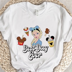 May include: White t-shirt with a graphic of a Disney princess with blonde hair and blue eyes. The princess is surrounded by Mickey Mouse ears and various Disney snacks. The text "Best Day Ever" is written in a whimsical font below the graphic.