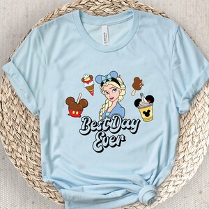 May include: Pale blue t-shirt with a cartoon of Elsa, ice cream, and the text "Best Day Ever". The shirt has rolled-up sleeves. The design includes various ice cream treats and a Mickey Mouse-shaped ice cream.