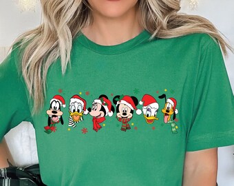 Op de afbeelding: Groen T-shirt met een kerstthema ontwerp met Mickey Mouse, Minnie Mouse, Donald Duck en Goofy die kerstmanmutsen dragen.