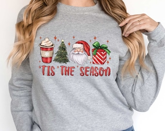 Op de afbeelding: Grijs sweatshirt met een kerstthema ontwerp met een kopje koffie, een kerstboom, de kerstman en een cadeaudoos. De tekst "'Tis the Season" is onder het ontwerp gedrukt.