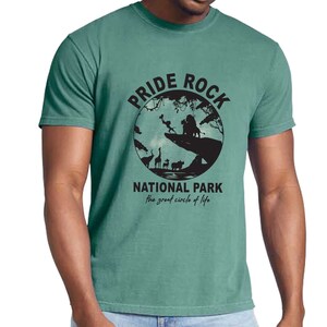 May include: A green t-shirt with a graphic of a circle with the words "Pride Rock National Park" and "the great circle of life" printed on it. The circle features a silhouette of a lion, a lioness, and other animals from the movie The Lion King.